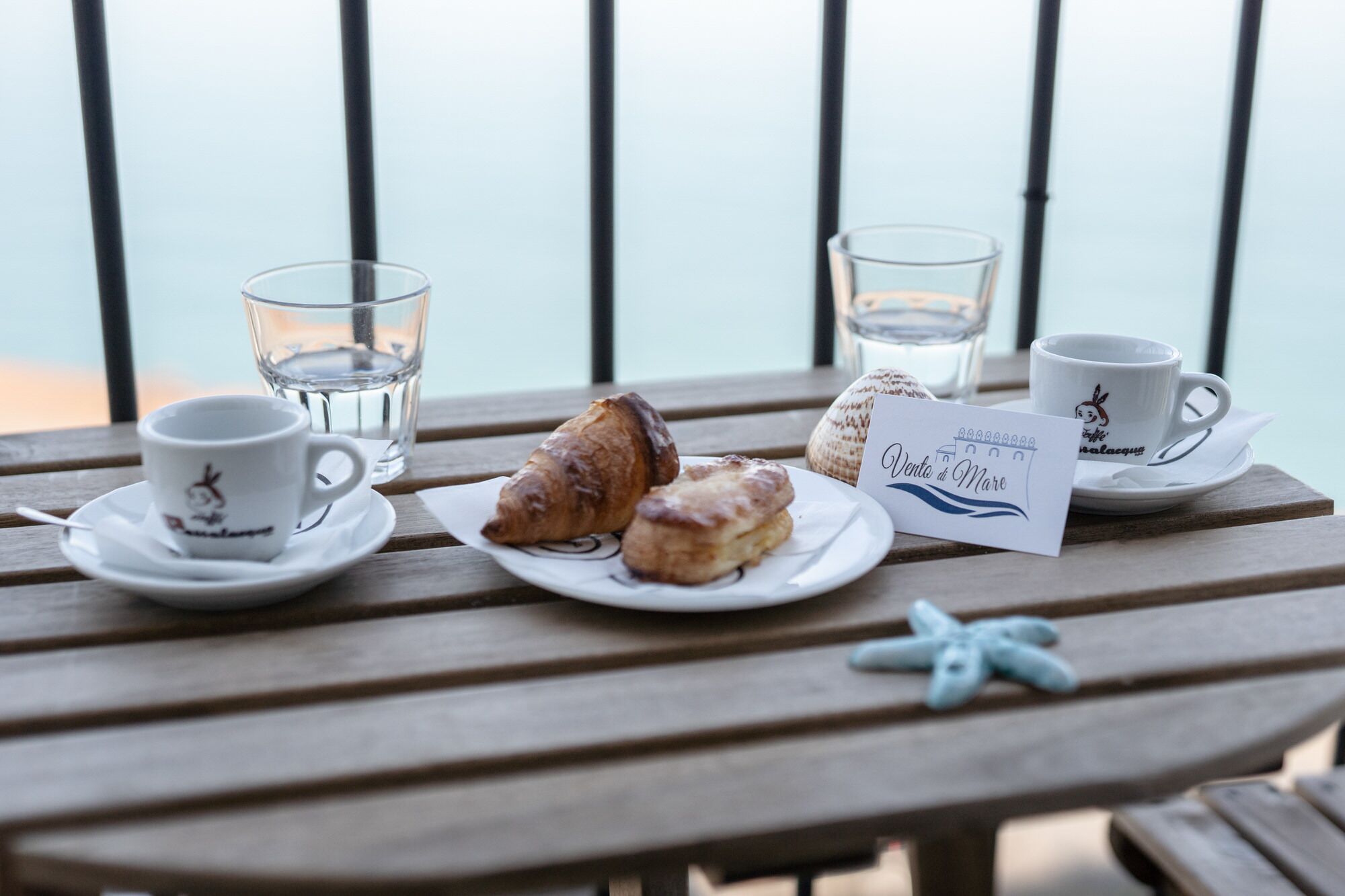Una mesa de madera con un plato de pasteles, dos tazas, un vaso de agua, una decoración de estrella de mar y un letrero que dice "Vento di Mare".
