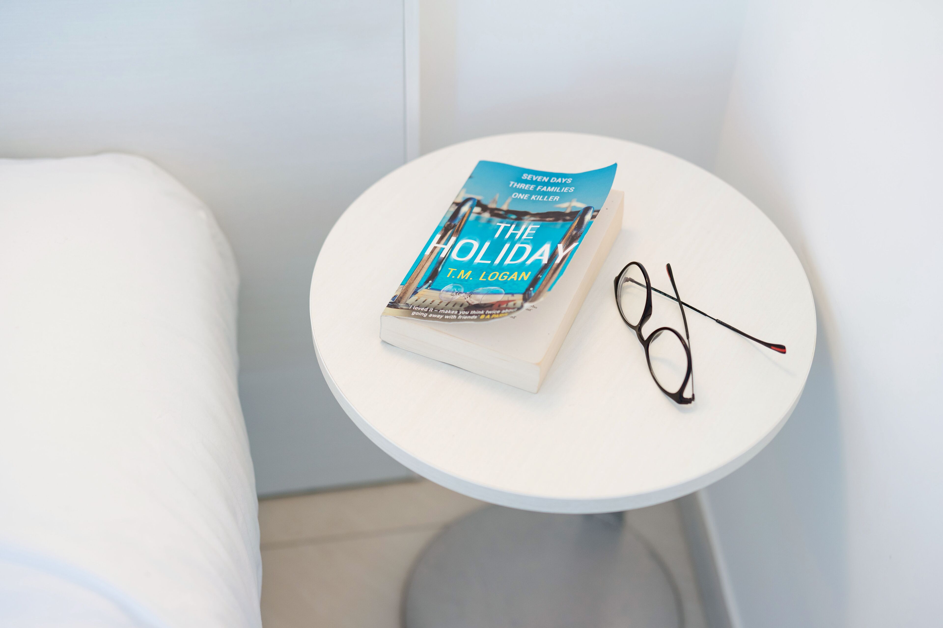 A book titled "The Holiday" by T.M. Logan on a white round table next to a white toilet.