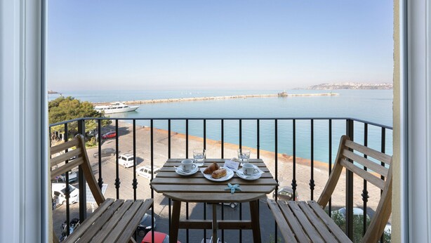 Un balcón con una mesa lista para dos personas, con vistas al mar tranquilo y a una costa lejana.