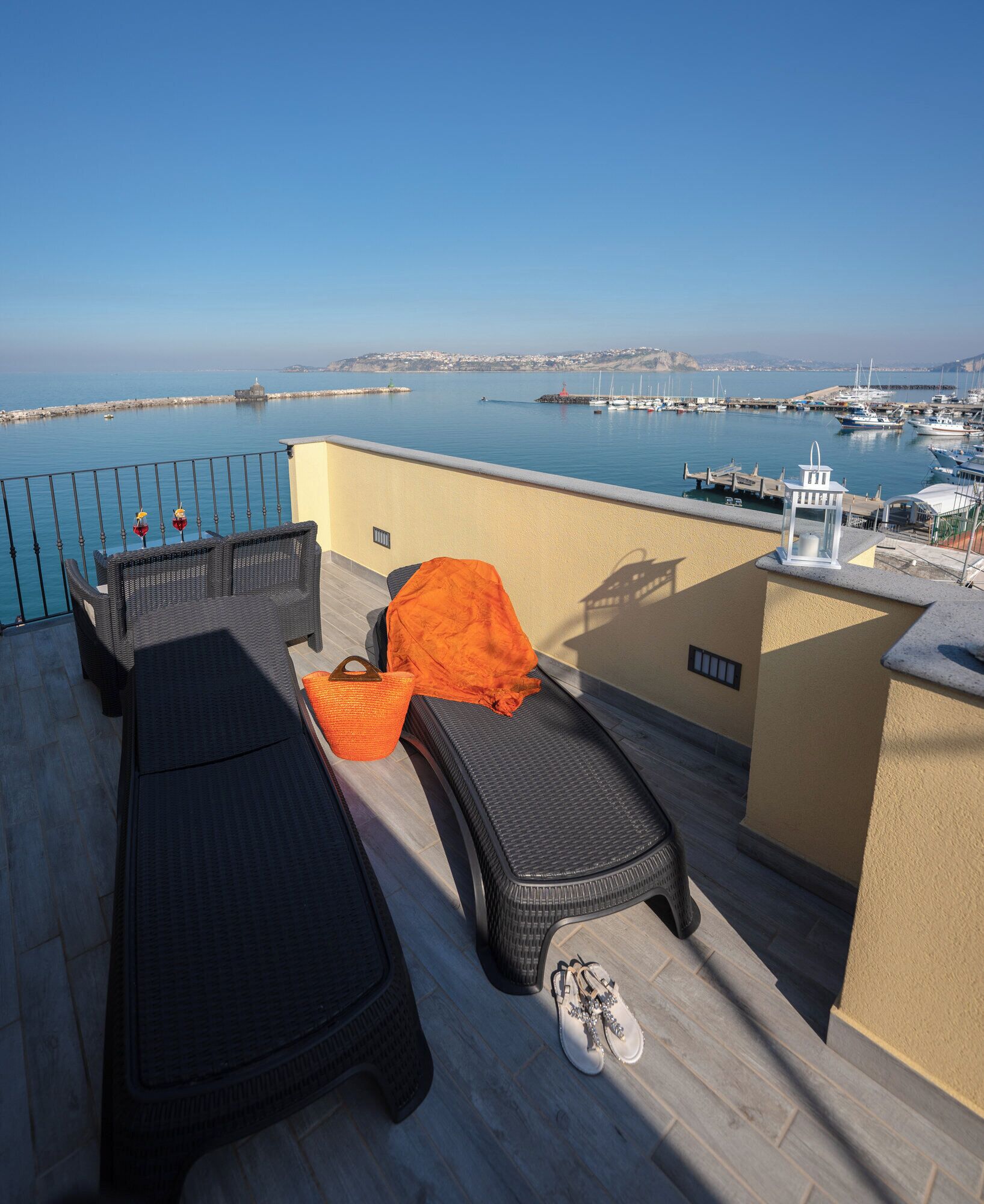 Une terrasse sur le toit, avec deux chaises longues, un petit coussin orange et une paire de chaussures. La terrasse offre une vue sur une marina avec des bateaux et une côte lointaine.