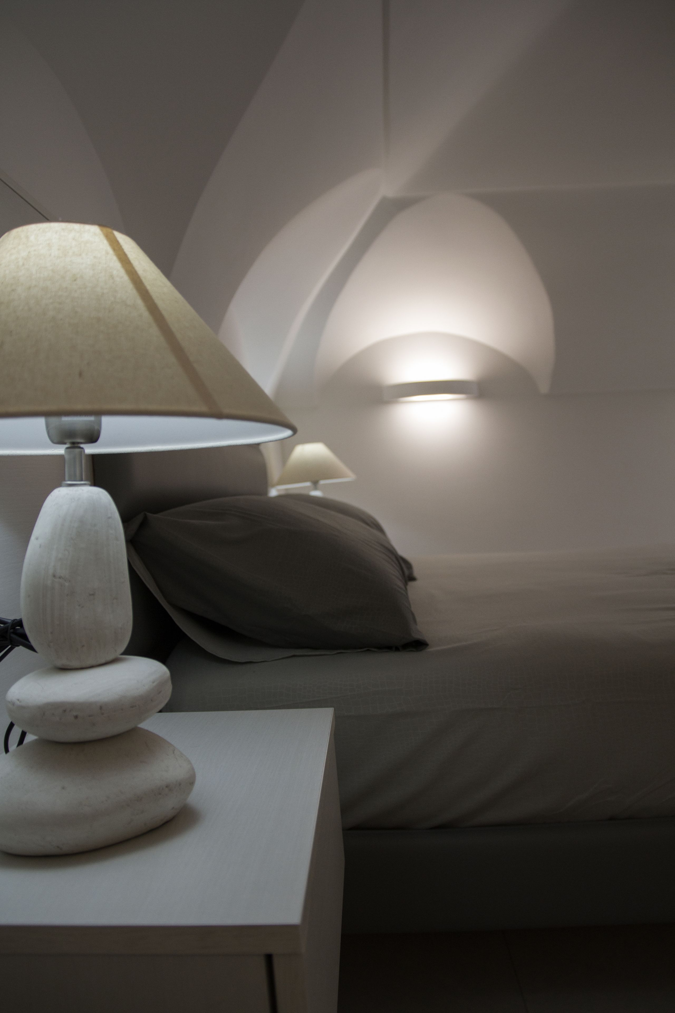 A bedroom with a bed, a nightstand with a lamp, and a wall-mounted light fixture.