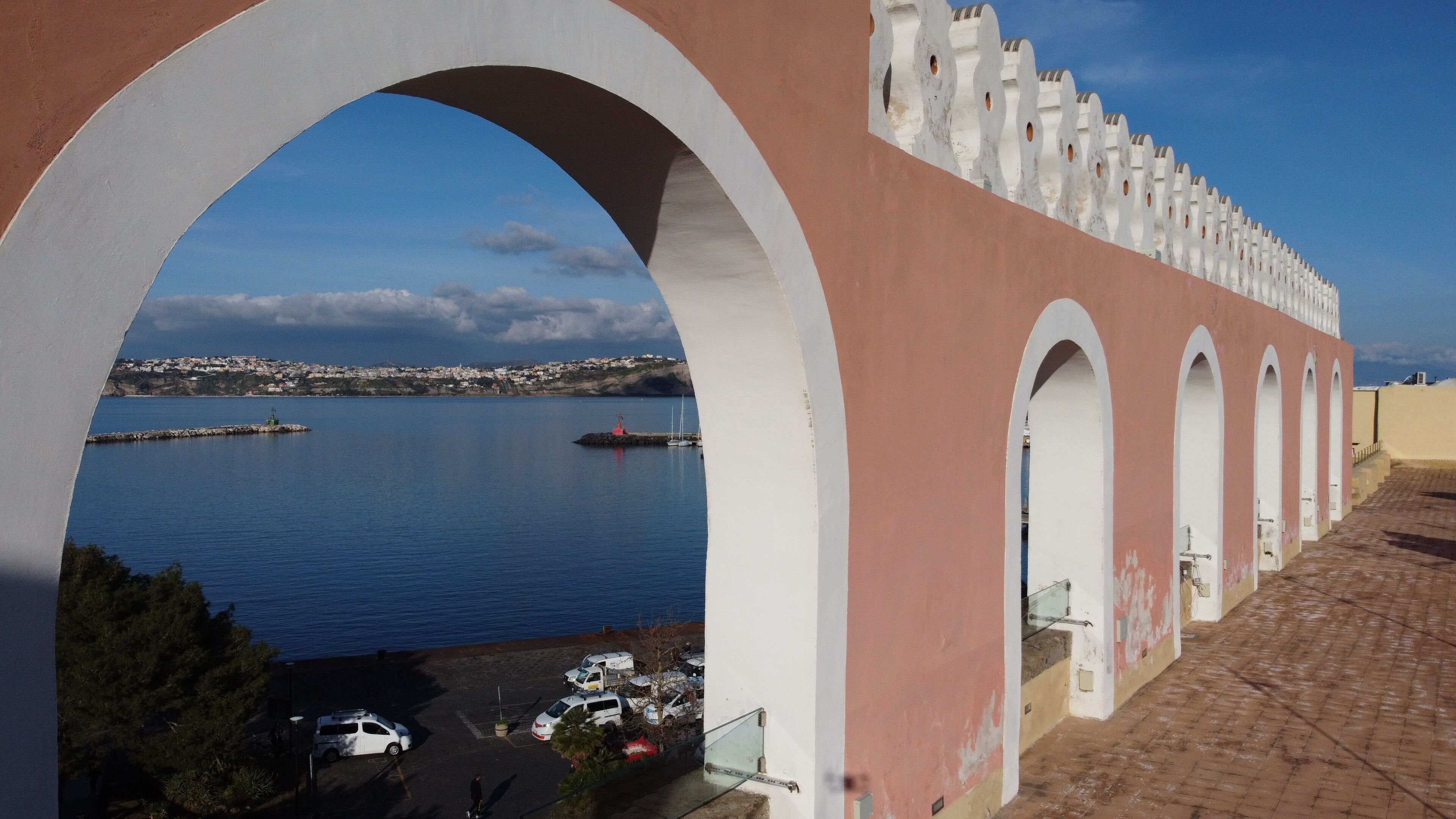 A coastal view through a series of arches overlooking a harbor with boats and a distant town.