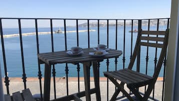 Un balcón con una mesa y una silla, con vista a una zona costera y al mar, con tierra a lo lejos.