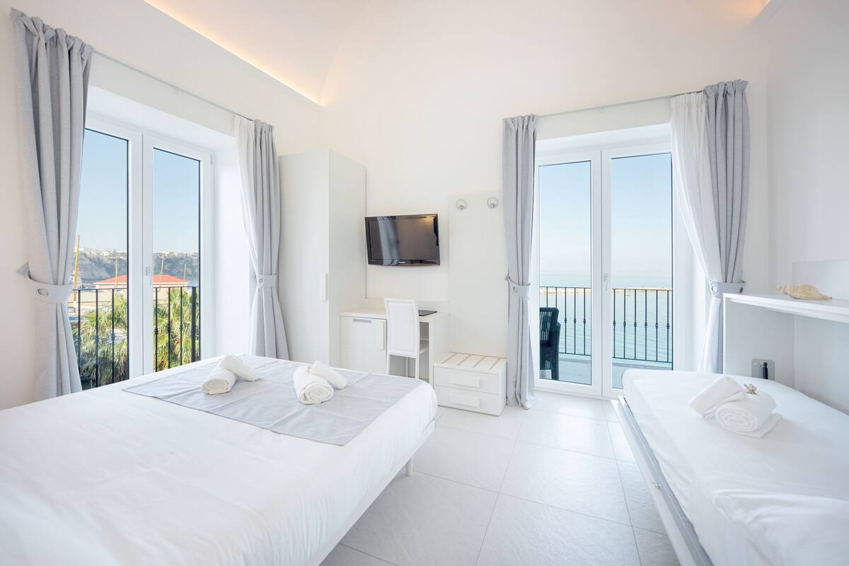 Habitación de hotel con dos camas, televisión, escritorio y balcón con vistas.