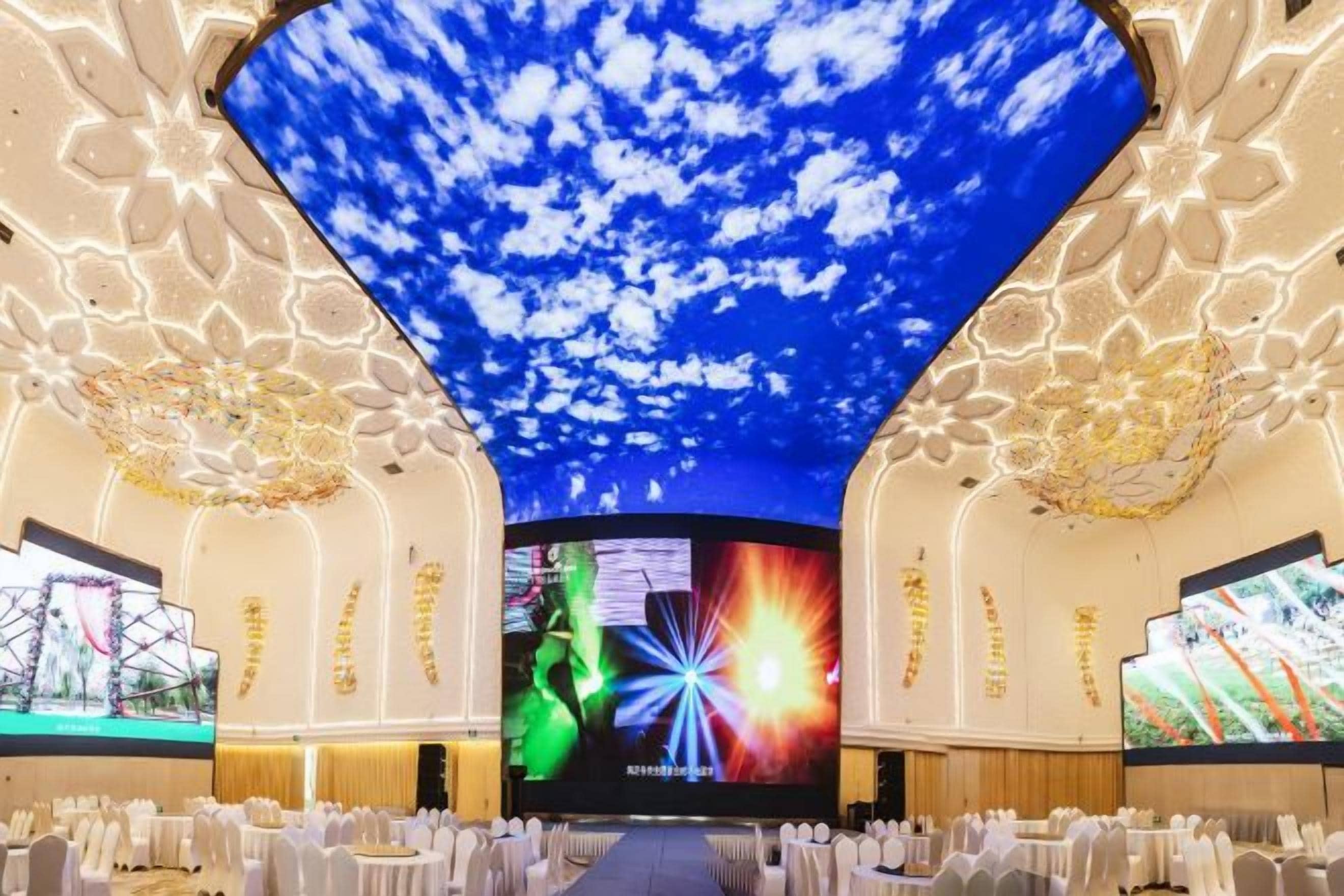 A conference room with a large ceiling screen displaying a sky with clouds, a central stage with a large screen, and rows of white chairs.