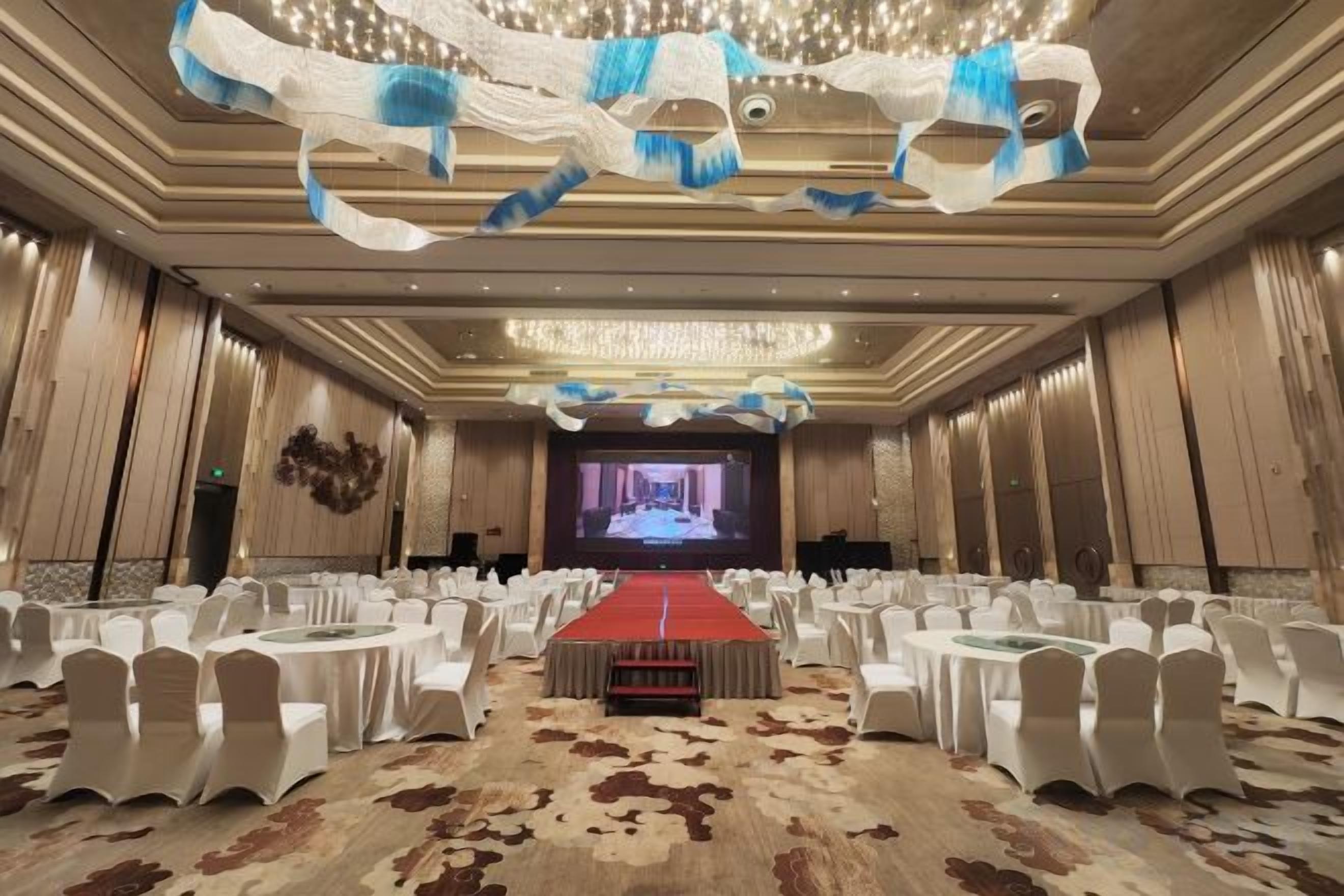 Un salón de conferencias con alfombra roja, sillas blancas y una pantalla grande al frente.