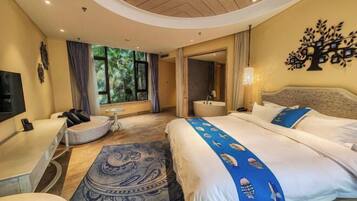 Habitación de hotel con cama, escritorio, silla, mesa redonda, espejo, lavamanos y ventana con vista a follaje.