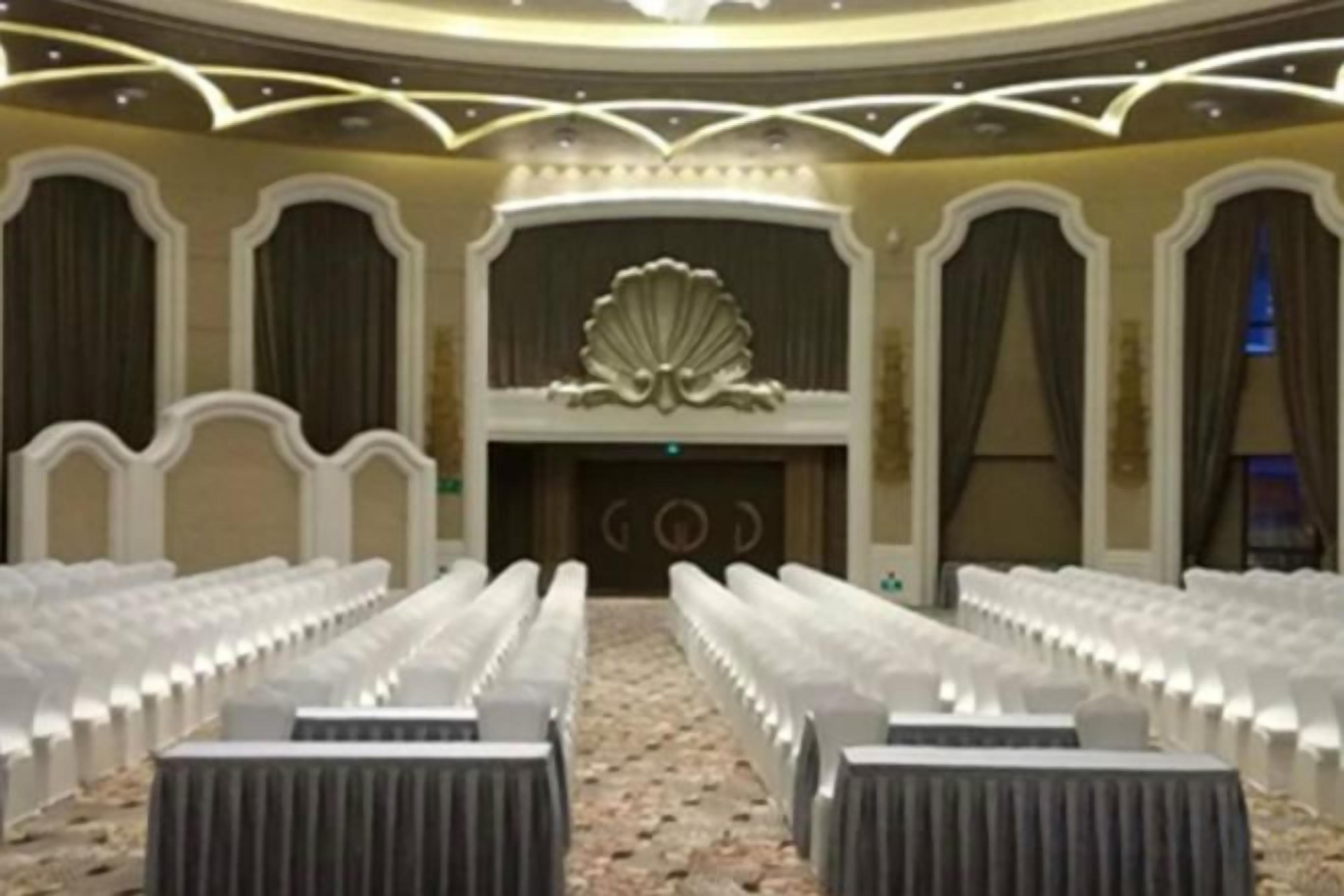 A conference room with rows of white chairs, a stage with a decorative backdrop, and a chandelier.