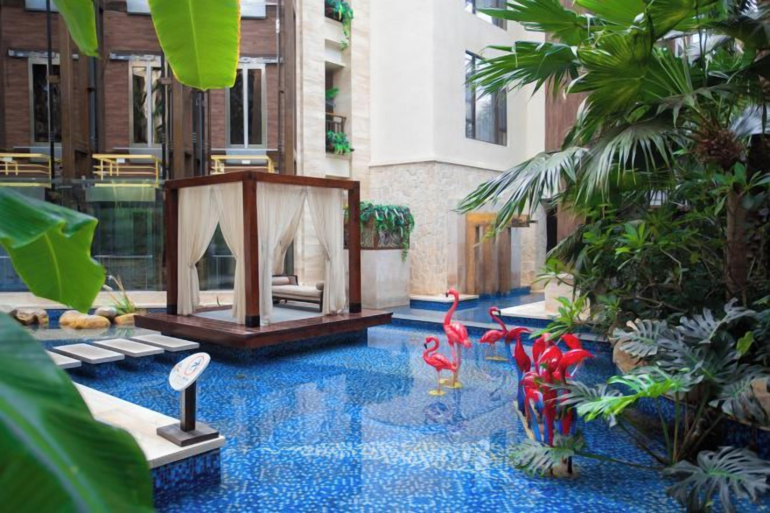 A pool area with a glass-enclosed seating area, surrounded by tropical plants and a decorative flamingo statue.