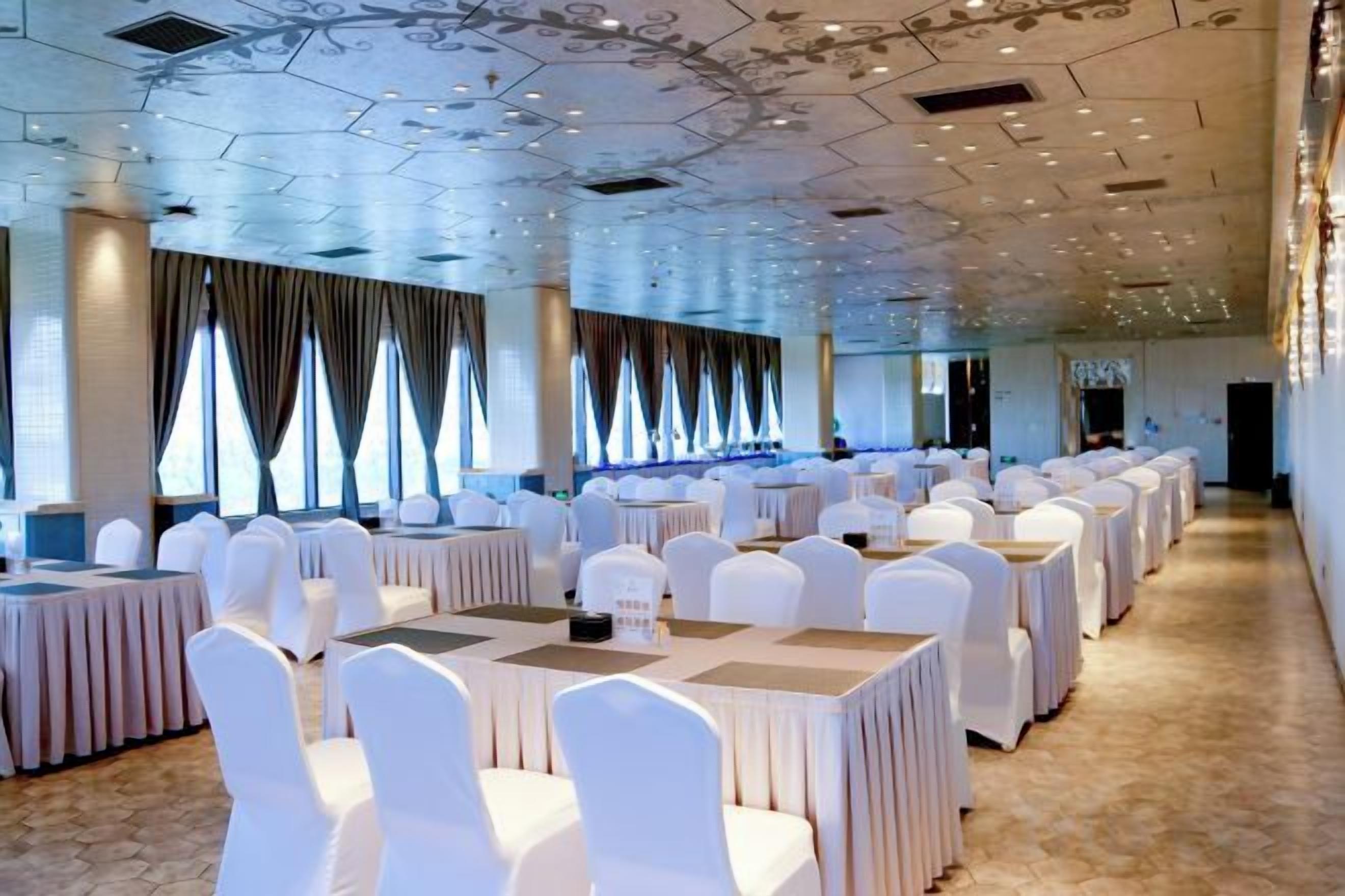 Une salle de banquet avec des tables rondes et des chaises blanches, de grandes fenêtres avec des rideaux et un plafond orné de motifs.