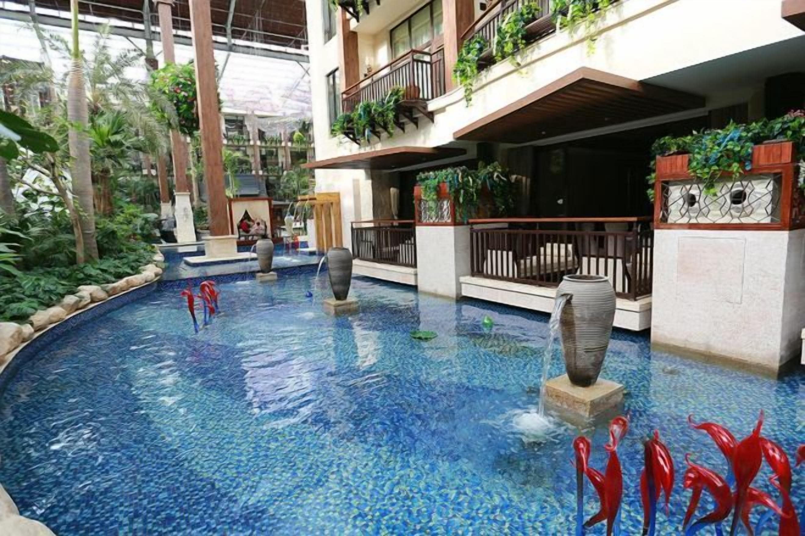 An indoor swimming pool with a fountain, surrounded by balconies with plants, and a view of the sky through the roof.