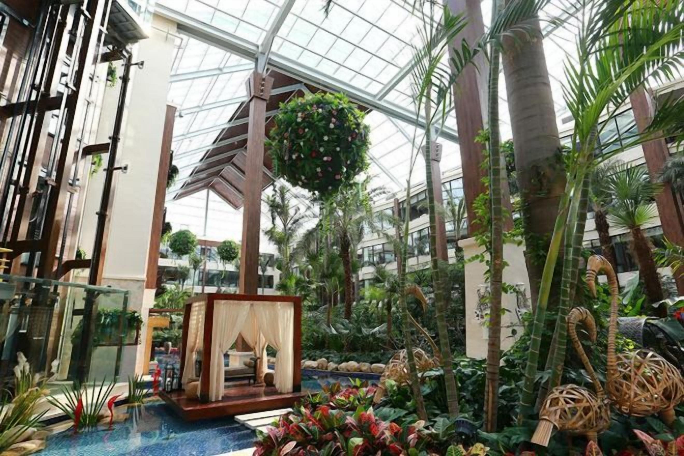 An indoor atrium with a glass ceiling, tropical plants, a seating area with a draped canopy, and a hanging flower arrangement.