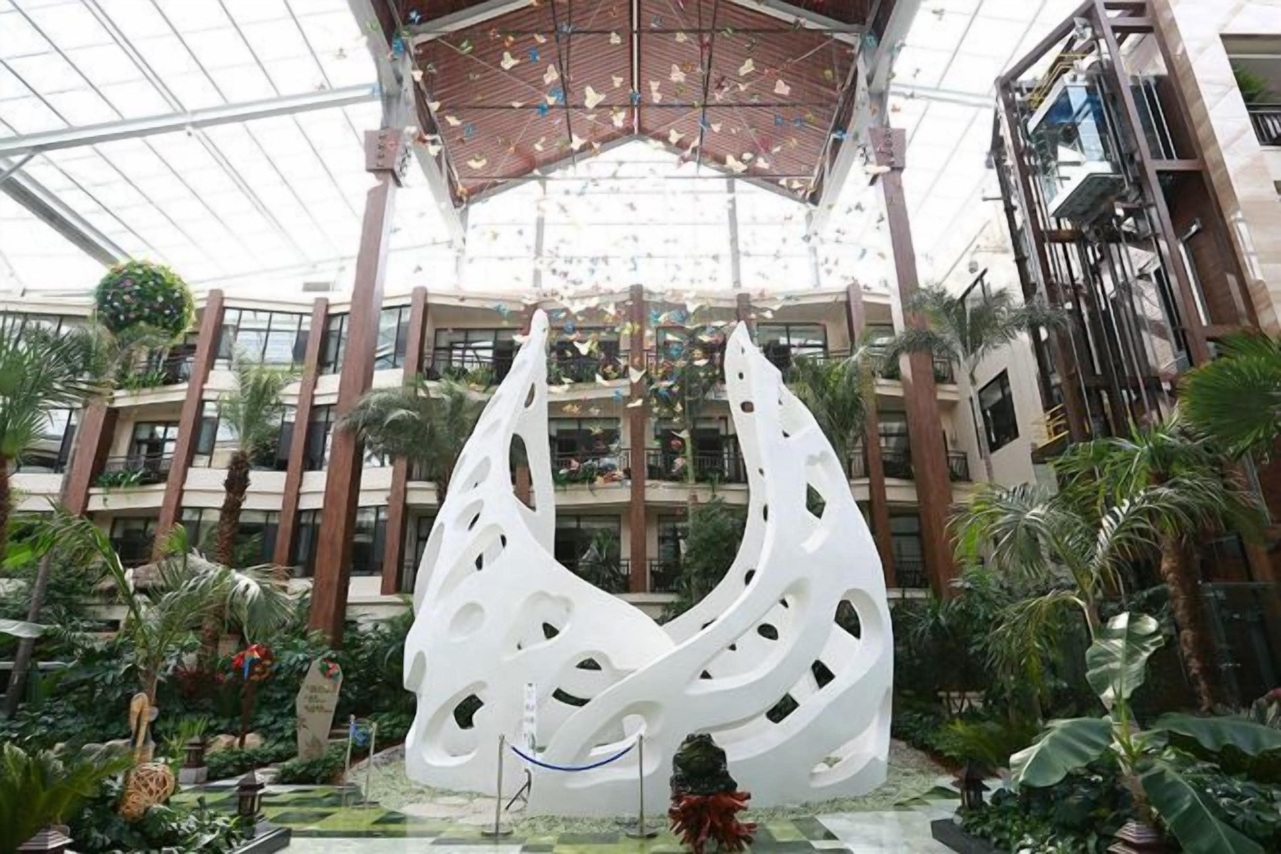A multi-level atrium with a central white sculpture, surrounded by indoor plants and a glass ceiling.