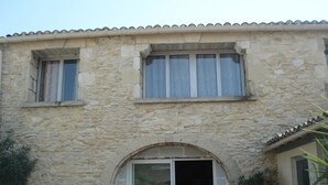 A stone building with a tiled roof, a central arched doorway, and two windows with curtains. - LA GARIGUETTE APARTMENT NEAR NIMES (Uchaud)