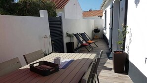 Una mesa de madera con sillas, una parrilla y una planta en maceta en un espacio al aire libre iluminado por el sol, con pared blanca y techo de baldosas.