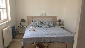 Un dormitorio con una cama, dos mesitas de noche, una lámpara, un radiador, una ventana y una cesta con la etiqueta "BAZAR".