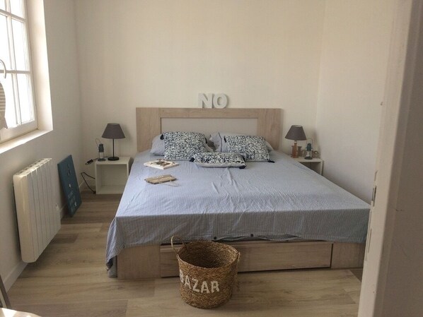 Een slaapkamer met een bed, twee nachtkastjes, een lamp, een radiator, een raam en een mandje met de tekst 'BAZAR'.