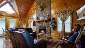 A cozy log cabin interior with a stone fireplace, leather seating, and a large window with curtains. - Close to Town-Outdoor Pool Access-Stocked Fishing Pond Access-private Putt Putt (Sevierville)