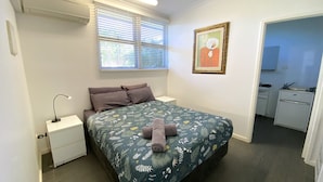 Un dormitorio con cama, mesita de noche, lámpara, ventana con persianas, un cuadro y un baño visible a través de una puerta abierta.