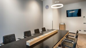 Kokoushuone, jossa on pitkä pöytä, tuoleja, kello, televisio ja kattovalaisimet.
