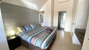 A modern bedroom with a striped bedspread, a nightstand with a lamp, and a view of the bathroom and kitchen through an open door. - Osprey Holiday Village Unit 203/1 Bedroom - Villa with a Pool in the Complex (EXMOUTH)