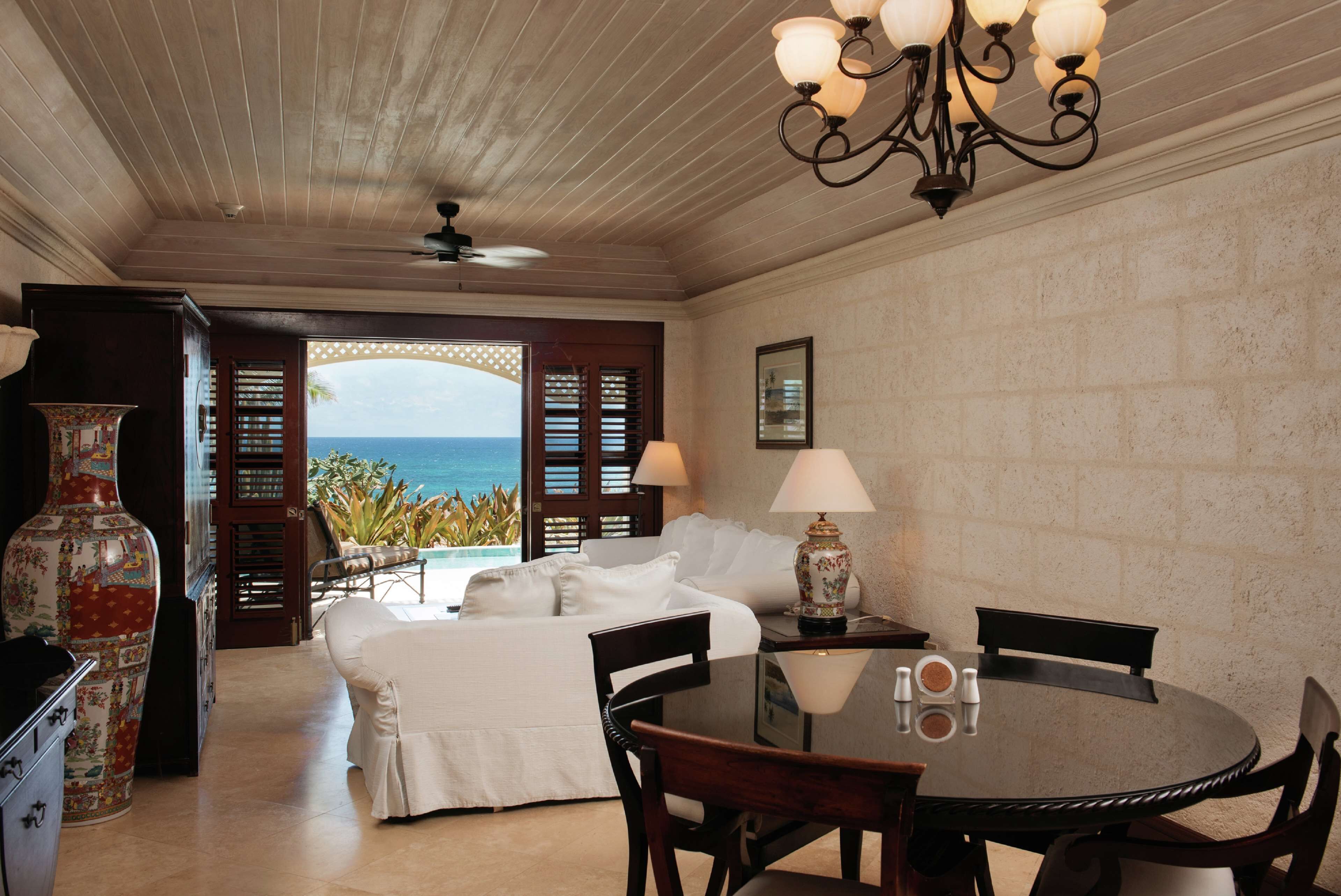 A room with a bed, a dining table, chairs, a chandelier, a vase, and a view of the ocean.