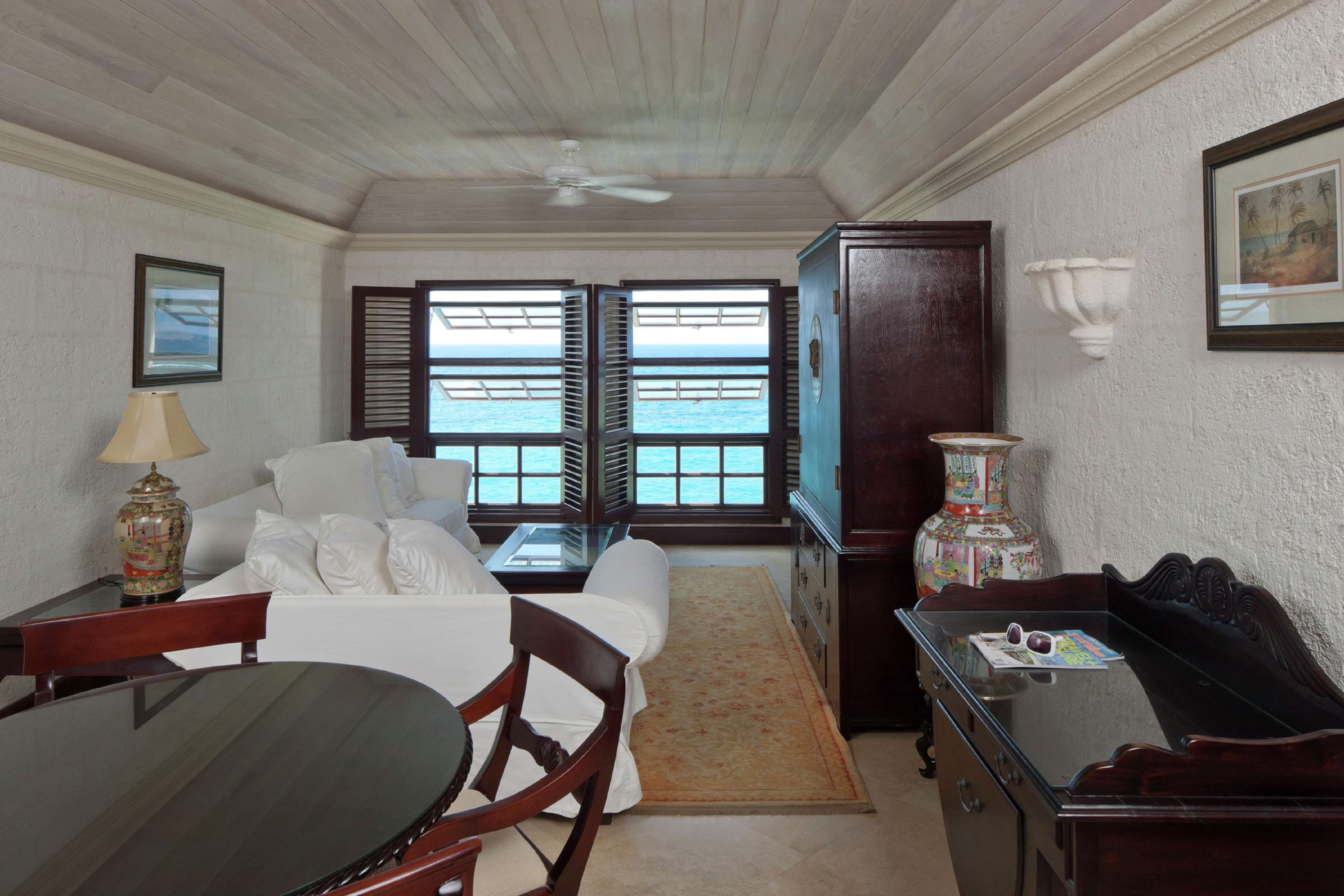 A room with a dining table, chairs, a sofa, a TV, and a view of the sea through open shutters.