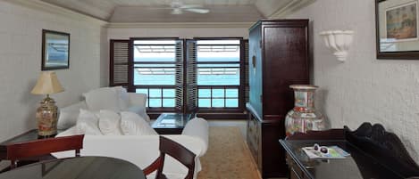 A room with a dining table, chairs, a sofa, a TV, and a view of the sea through open shutters.