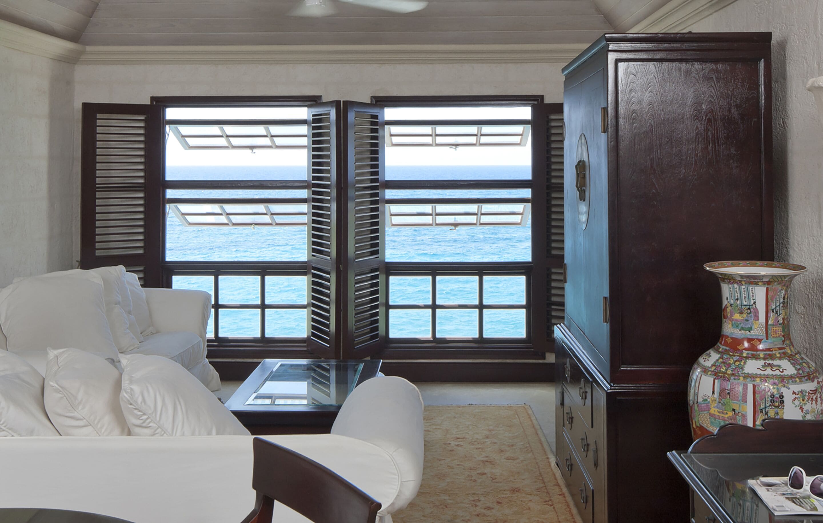 A room with a white sofa, a glass coffee table, a decorative vase, and a view of the ocean through open shutters.