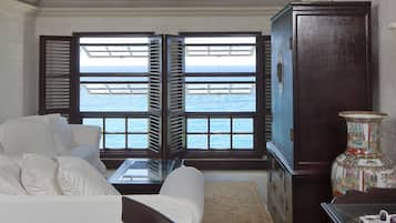 A room with a white sofa, a glass coffee table, a decorative vase, and a view of the ocean through open shutters.