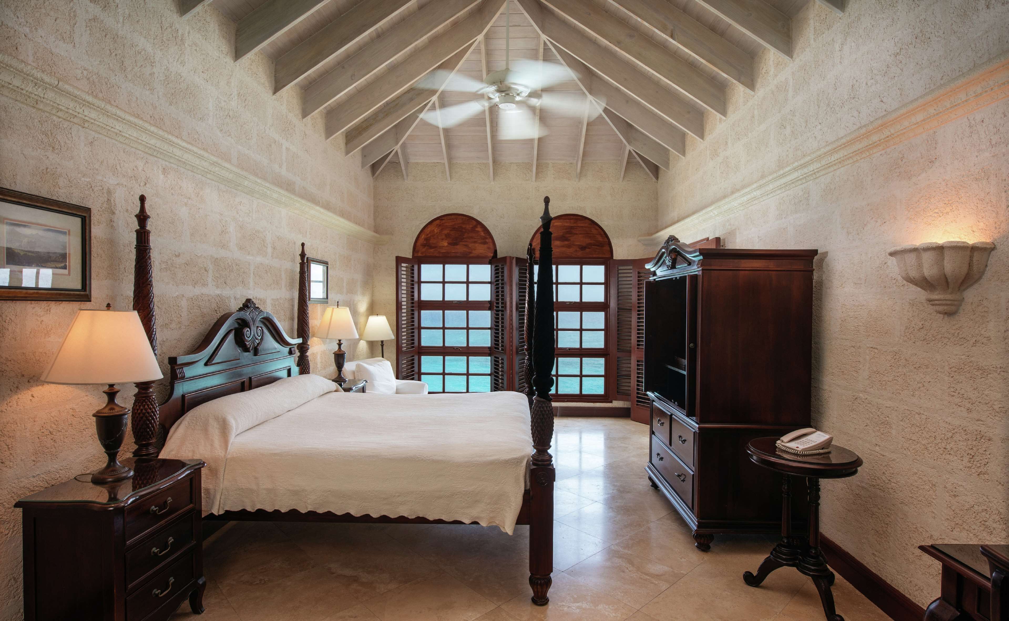 Un dormitorio amplio con una cama grande, un ventilador de techo y vistas al mar a través de ventanales arqueados.