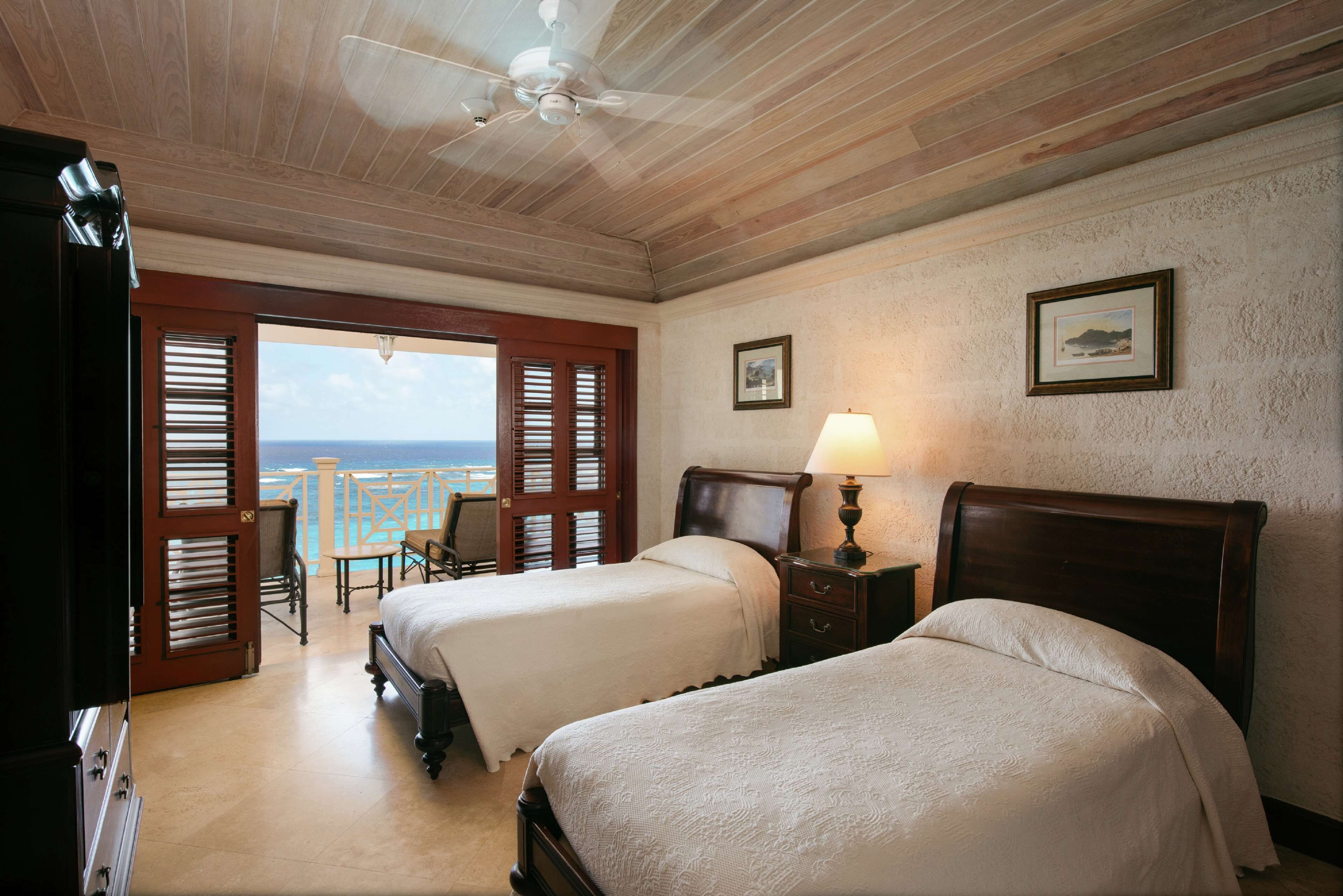 A hotel room with two beds, a ceiling fan, a view of the ocean, and a balcony with a table and chairs.