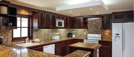 A modern kitchen with granite countertops, dark wooden cabinets, a built-in microwave, a stove, and a refrigerator.