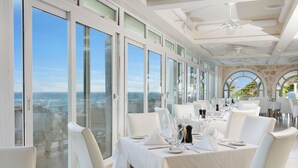 A dining area with white tables and chairs, large windows, and a view of the ocean.