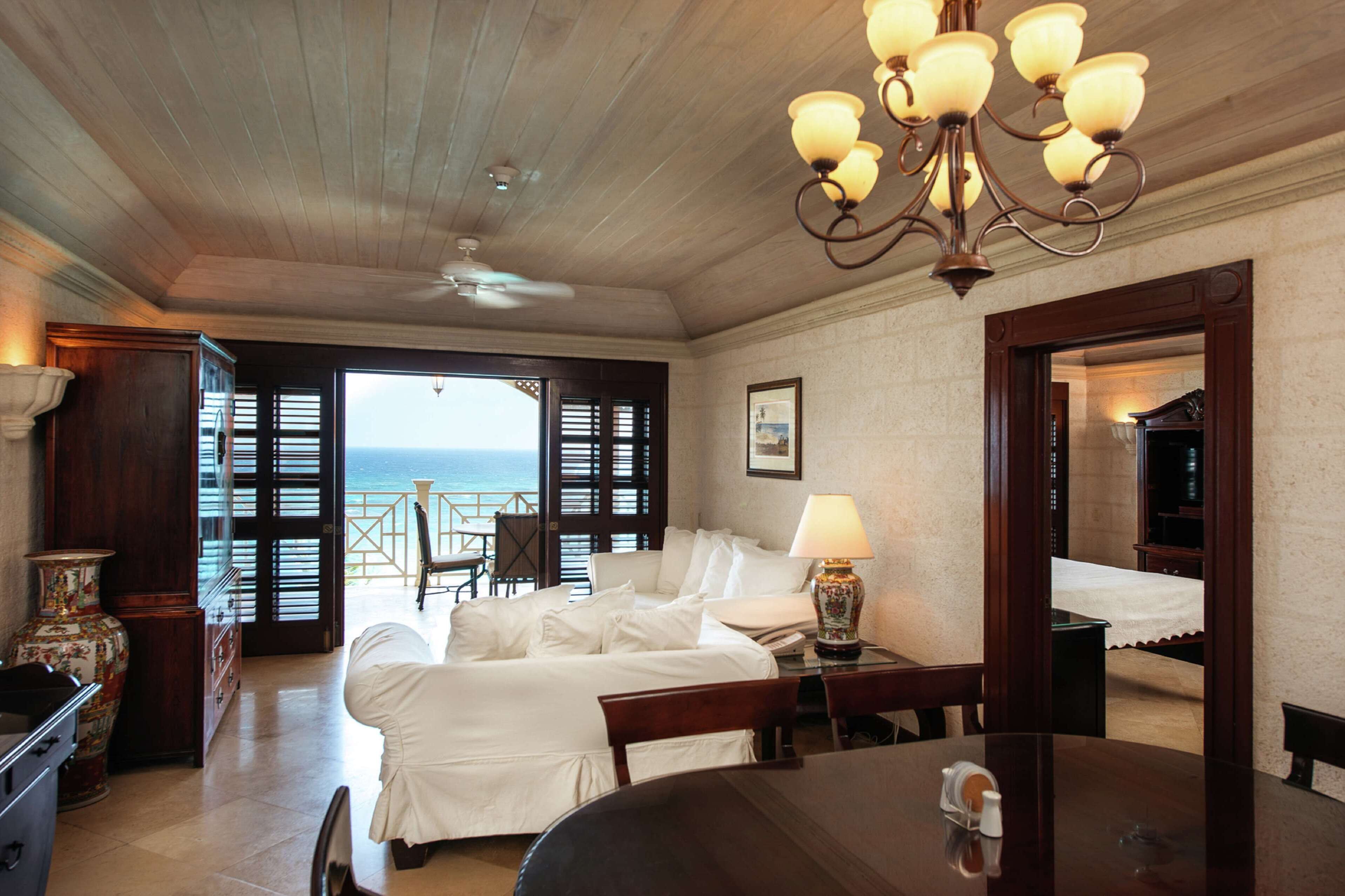 A hotel room with a bed, a dining table, a lamp, and a view of the ocean through the open door.