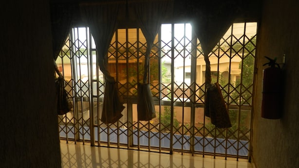 Una habitación con vistas a través de una ventana con barrotes, un extintor de incendios en la pared y el reflejo de una persona en el suelo brillante.