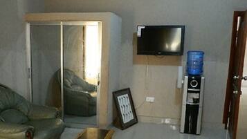 Una sala de estar con un sofá, un televisor montado en la pared, un dispensador de agua y una puerta que da a otra habitación.