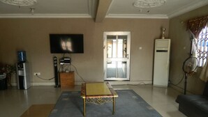 Una sala de estar con un televisor de pantalla plana montado en la pared, una cafetera, un pequeño mueble de madera, una mesa con mantel estampado, un sofá y un ventilador de pie.