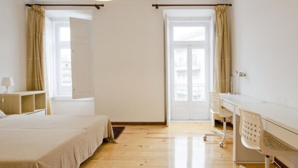Ein Schlafzimmer mit einem Bett, einem Schreibtisch und einem Sessel. Es gibt zwei Fenster mit Vorhängen.