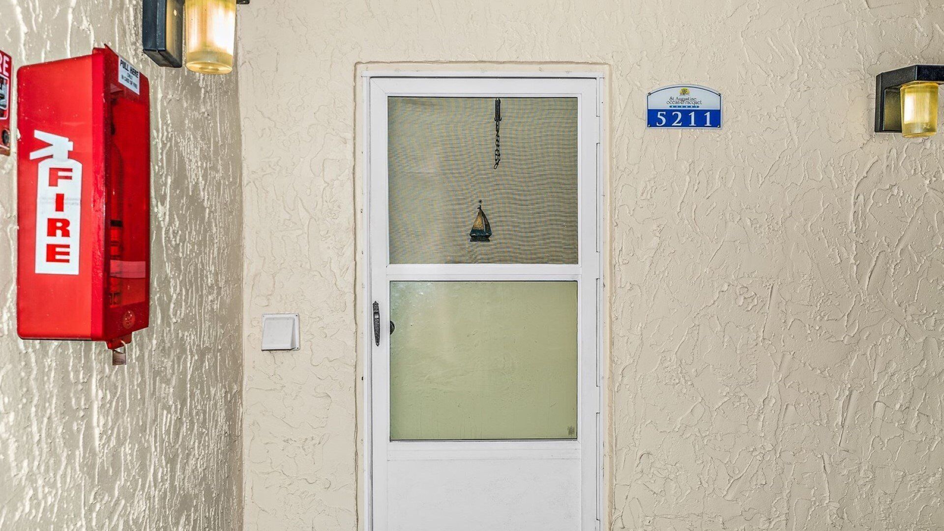 Entrada de um edifício com uma porta branca, um painel de alarme de incêndio e uma placa azul com o número 5211.