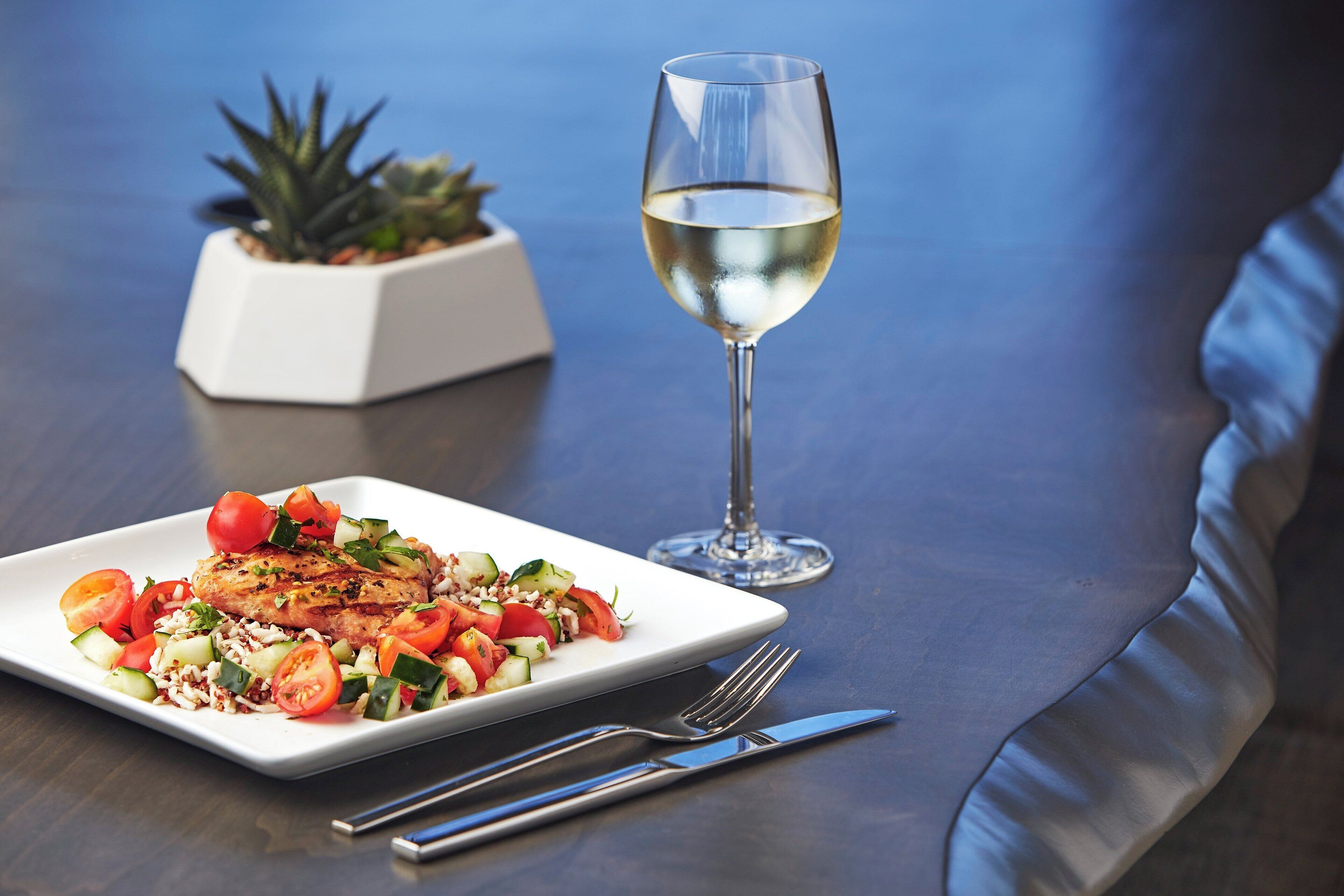 A dining table with a plate of food, a glass of white wine, and a small potted plant.