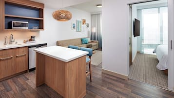 A modern hotel room with a kitchenette, a sofa, a dining area, and a large window.