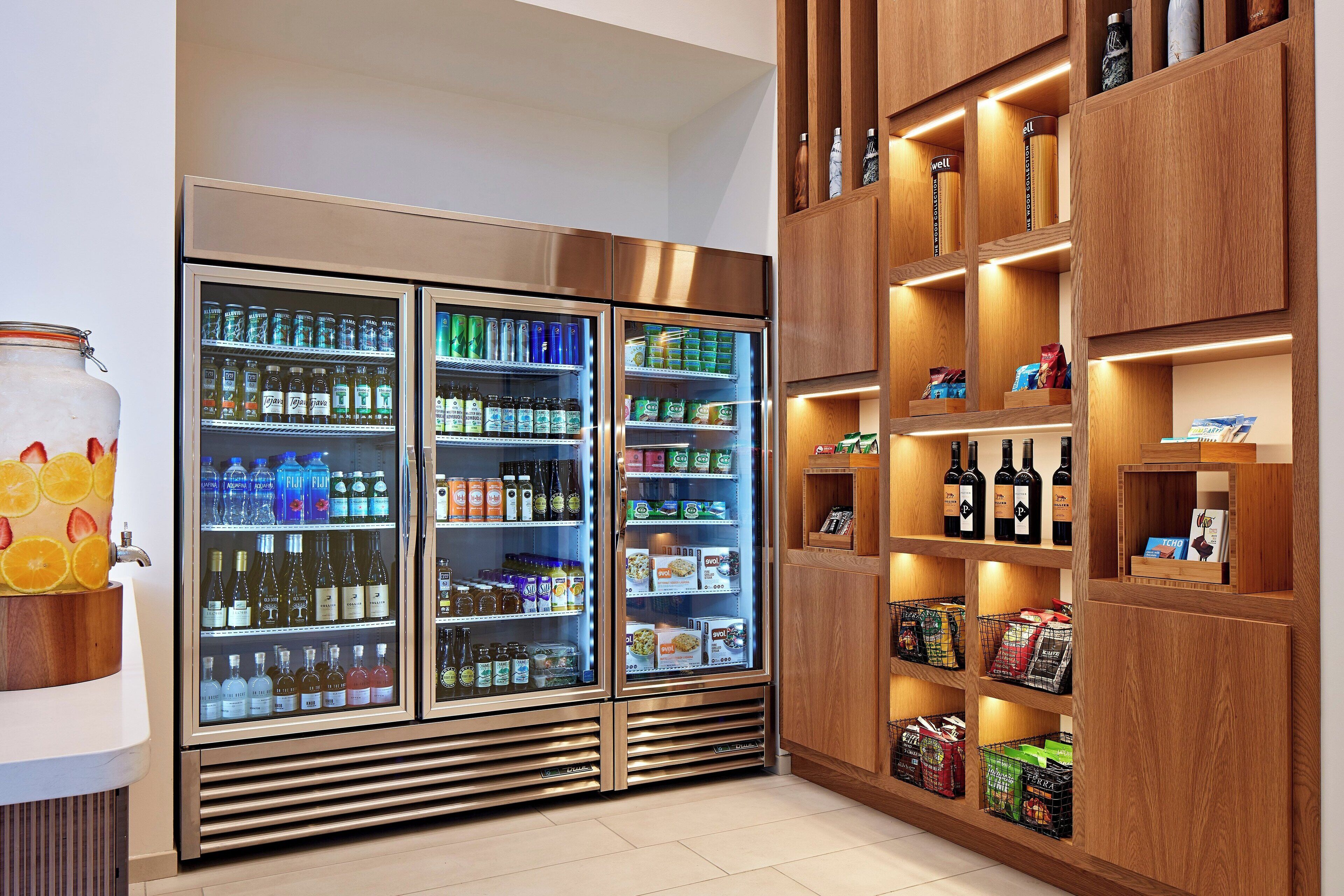 A refrigerated display with various beverages and snacks, wooden shelving with books and bottles, and a fruit bowl with oranges.
