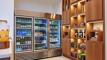 A refrigerated display with various beverages and snacks, wooden shelving with books and bottles, and a fruit bowl with oranges.