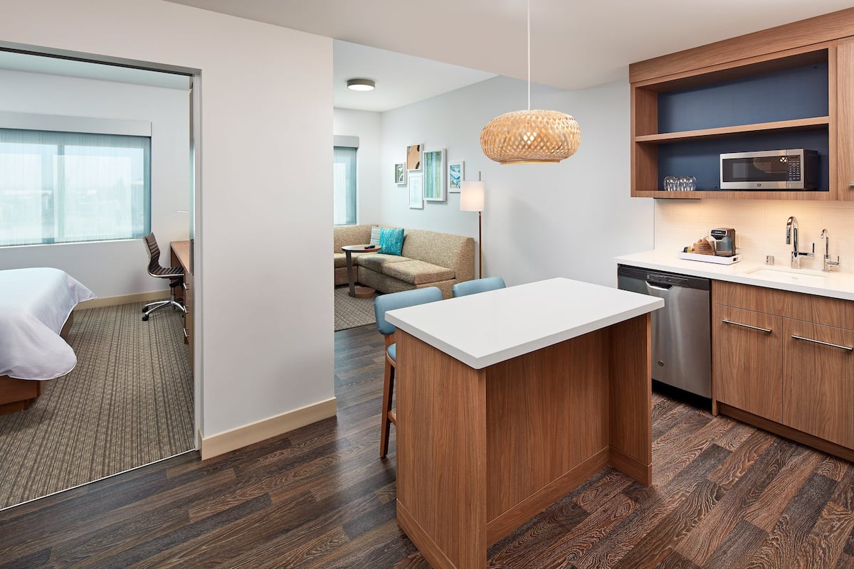 A modern hotel room with a wooden floor, a white bed, a kitchenette with a microwave, and a dining area with a sofa and chairs.