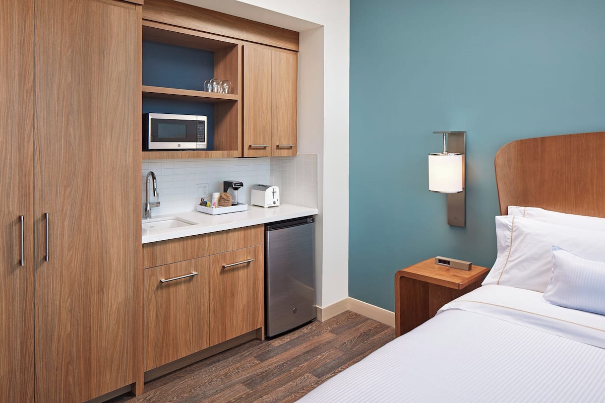 A hotel room with wooden cabinets, a microwave, a coffee maker, a bedside table with a lamp, and a bed with white linens.