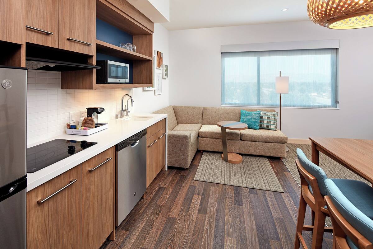 A modern kitchen with a built-in microwave, stainless steel refrigerator, and wooden cabinets. There is a small dining area with a table and chairs, and a large window providing a view of the outdoors.
