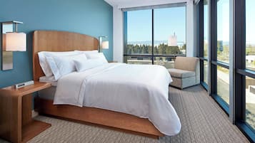 A modern hotel room with a large bed, a nightstand, a chair, and a view of the city through large windows.