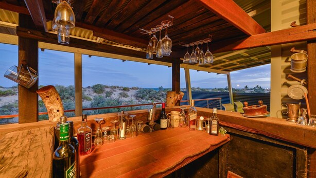 A bar area with a wooden counter, various bottles, glasses, and a view of the desert landscape through large windows.