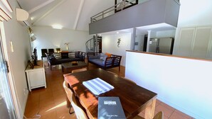 A spacious living area with a sofa, coffee table, and a dining table with a book titled "Exmouth Holidays." A staircase leads to an upper level. - Osprey Holiday Village Unit  112 - 3 Bedroom Fully Self-Contained Holiday Accommodation (EXMOUTH)